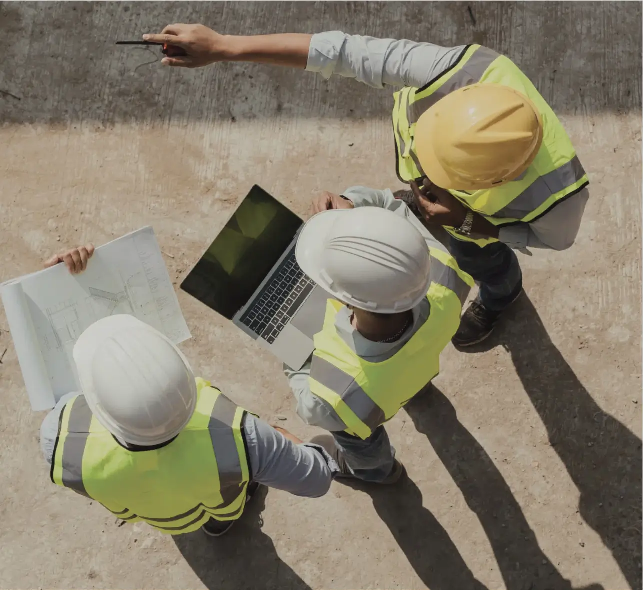 Three site workers reviewing plans and a laptop on a construction site